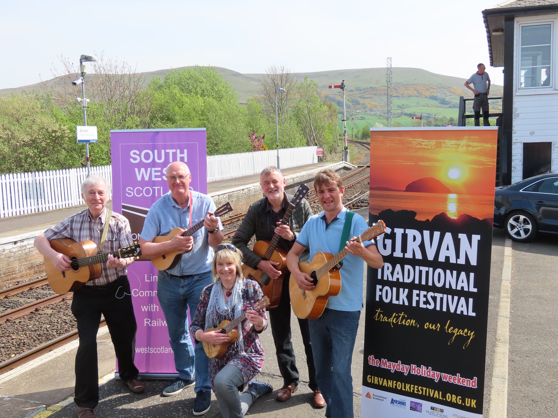 A group of folk musicians stand, smiling and holding instruments, by a railway line in front of banners promoting the Girvan Traditional Folk Festival